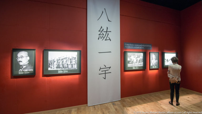 Visitor viewing framed historical photographs beside a tall white panel of Japanese characters on a red gallery wall.