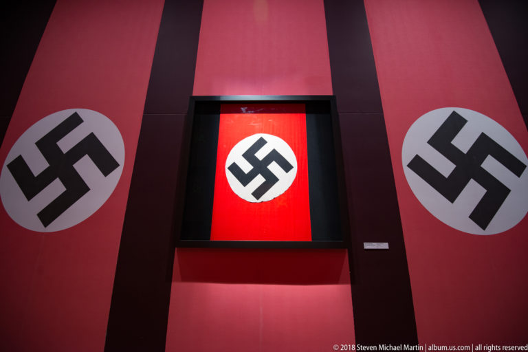 Red museum wall with large black swastikas in white circles surrounding an illuminated central swastika banner.