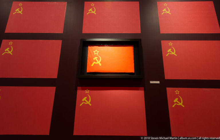 Red gallery wall covered with repeated yellow hammer-and-sickle symbols around a brightly lit central panel.