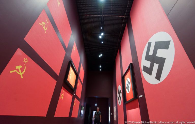 Angled red museum walls displaying black swastikas on one side and yellow hammer-and-sickle emblems on the other.