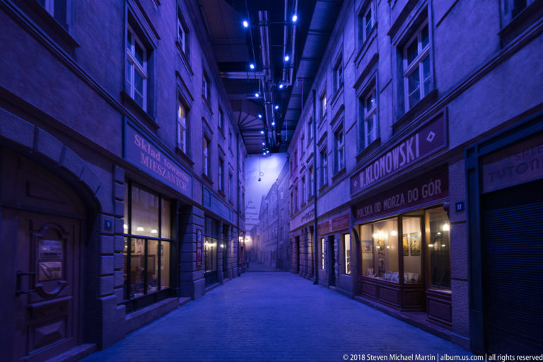 Blue-lit reconstructed city street with Polish shop signs, apartment façades, and illuminated storefronts inside the museum.