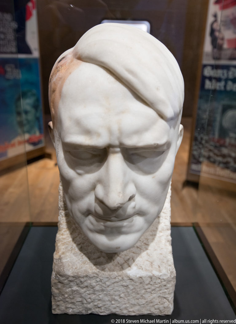 White stylized sculpted male head with downcast eyes mounted on a rough rectangular base in a museum case.