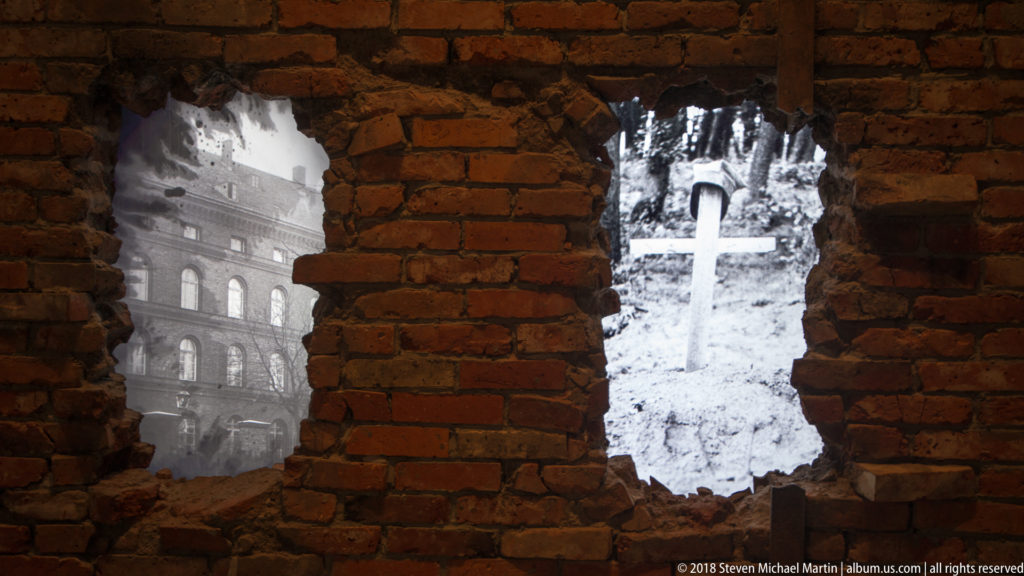 Two jagged openings in a red brick wall containing black-and-white projections of wartime destruction and a grave marker.