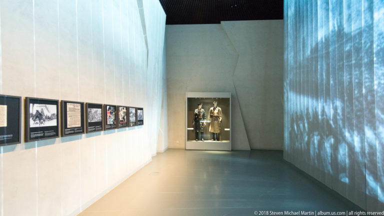Long museum corridor with framed displays at left, blue historical projection at right, and uniformed figures ahead.