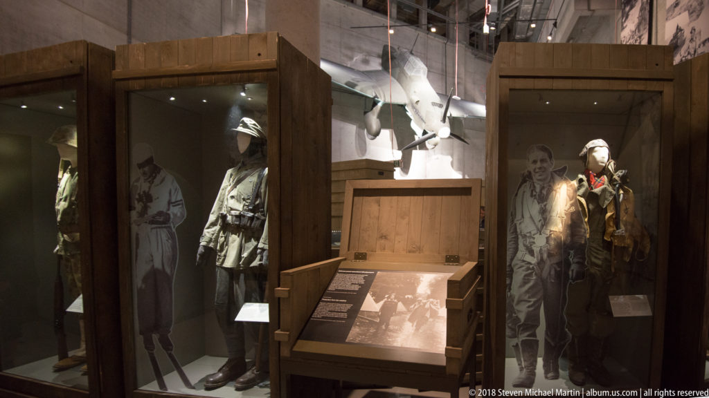 Military uniforms on mannequins in wooden cases with a suspended propeller aircraft visible behind them.