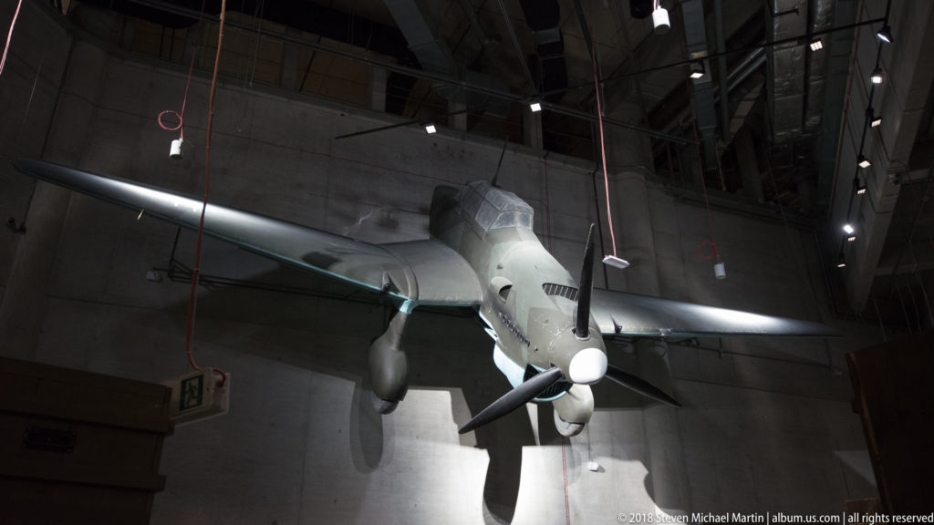 Single-engine propeller aircraft suspended nose-down slightly in a tall concrete museum gallery.