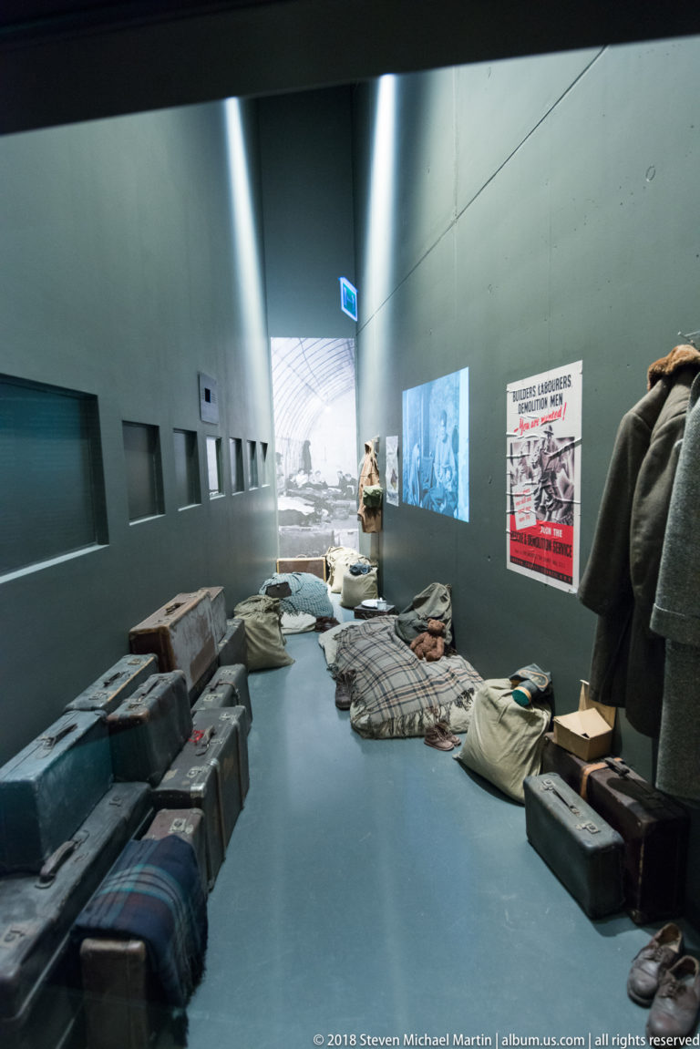 Suitcases, blankets, clothing, bedding, and bundled belongings arranged along a gray museum gallery.