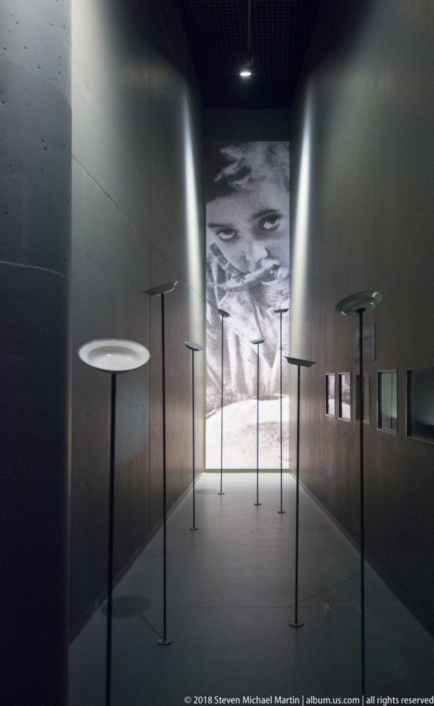 Dark narrow museum corridor with thin pedestal stands and a large black-and-white face at the far end.