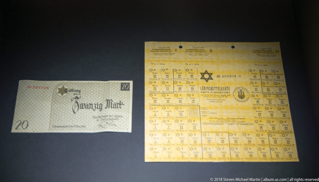 Small 20-mark paper note beside a yellow Star of David document with a printed grid in a museum case.
