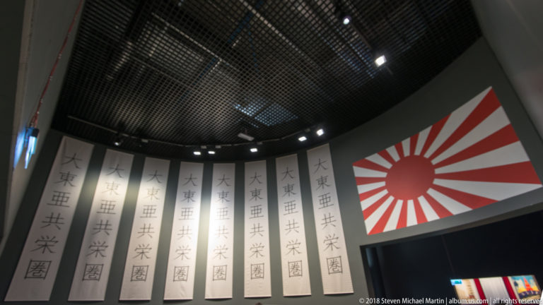 Japanese Rising Sun flag beside tall white banners with East Asian characters in a dark museum gallery.