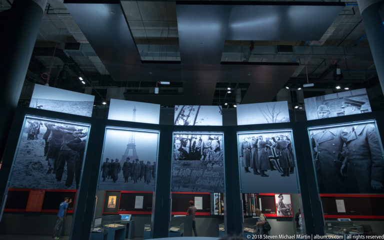 Dark gallery with enlarged black-and-white wartime photographs above artifact cases and red-accented labels.
