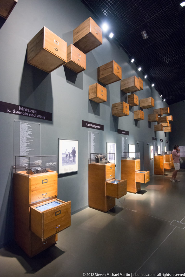Open wooden drawers and luggage-like cases arranged along a museum gallery with photographs and bilingual wall text.