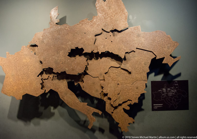 Large tan relief map with internal boundary lines and cutout gaps mounted on a gray museum wall.