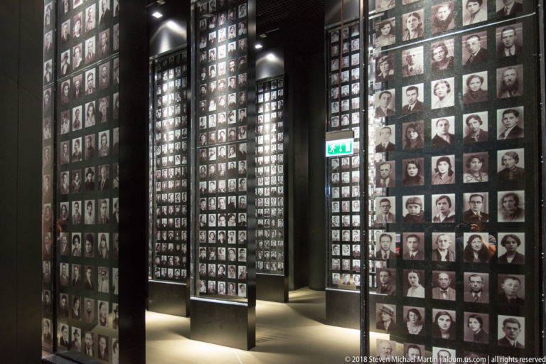Multiple tall panels filled with black-and-white identification portraits receding through a museum gallery.