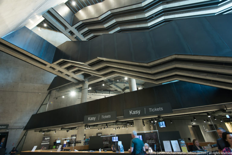 Angular dark ceiling planes above bilingual Kasy/Tickets counters with visitors in the museum lobby.