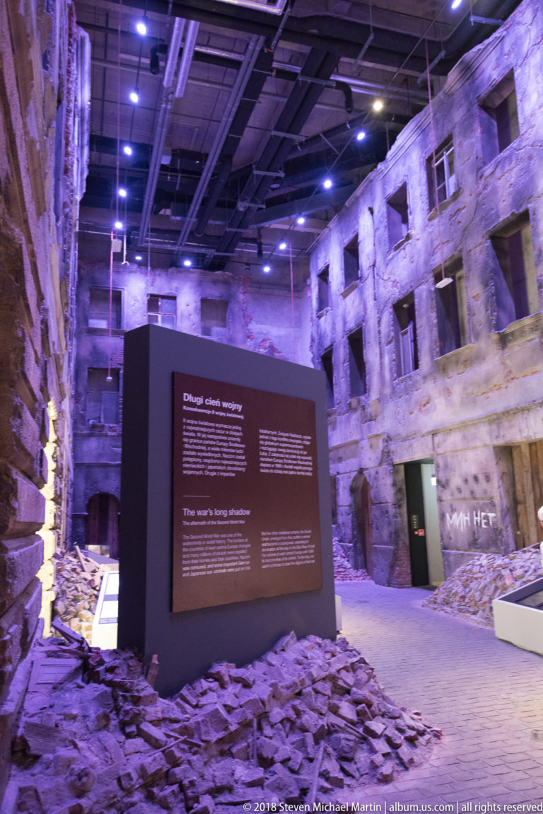 Interpretive panel surrounded by rubble and reconstructed damaged façades in a dark museum gallery.