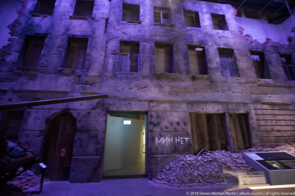 Reconstructed damaged building façade with broken windows, rubble, boarded doors, and purple exhibition lighting.