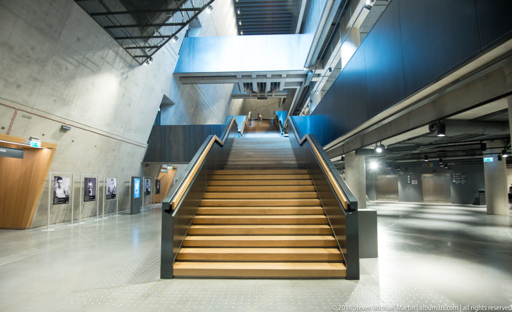 Wide wooden staircase rising through a tall concrete museum atrium with open galleries on both sides.