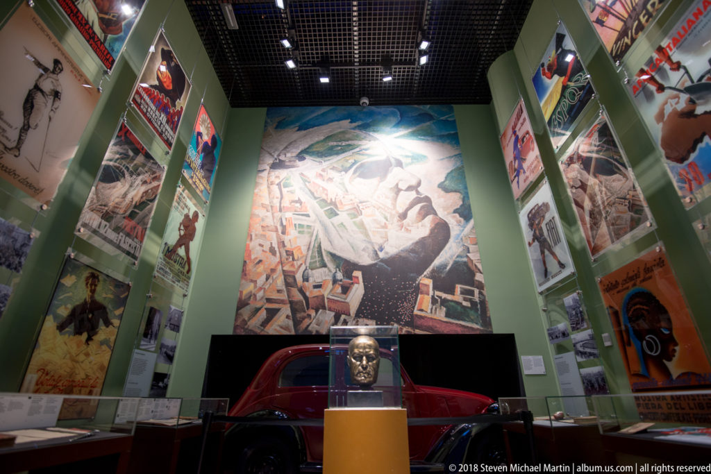 Gallery with towering political mural, propaganda posters on both walls, red automobile, and sculpted head in foreground.