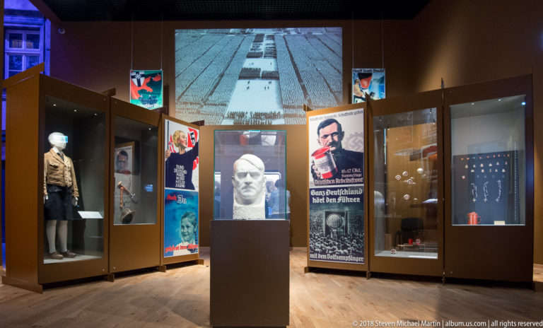 Central white sculpted head on a pedestal surrounded by propaganda posters, uniform displays, and a projected crowd scene.