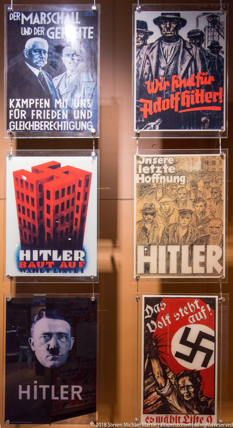 Glass display filled with German propaganda posters featuring Adolf Hitler portraits, swastikas, slogans, and mass-rally graphics.