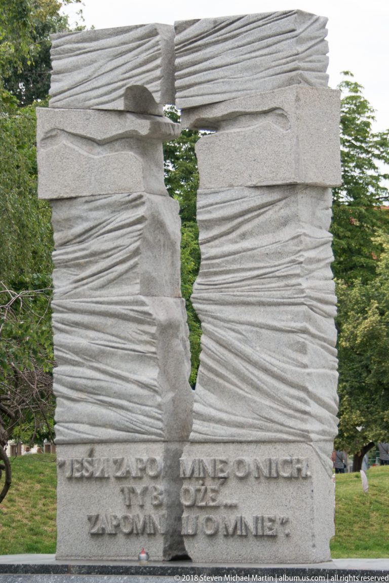 Split gray stone memorial with a vertical opening, horizontal carved grooves, and Polish inscription near the base.