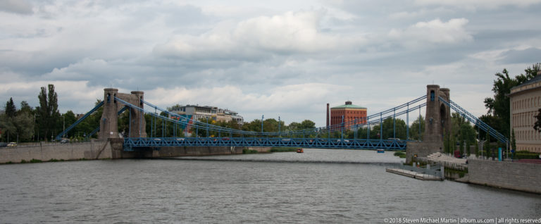 Blue suspension bridge with massive stone pylons spans the Oder River beneath a cloudy sky.