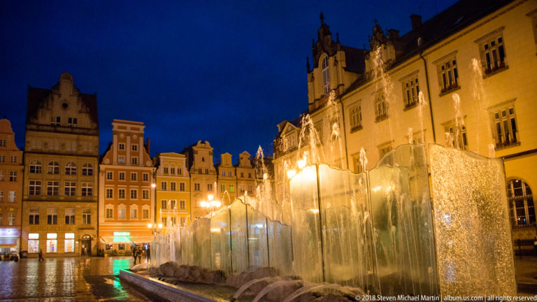 Illuminated glass fountain beside the Gothic Old Town Hall and historic façades on Wrocław Market Square.