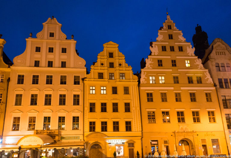 Three illuminated historic townhouses with curved and stepped gables on Wrocław Market Square.
