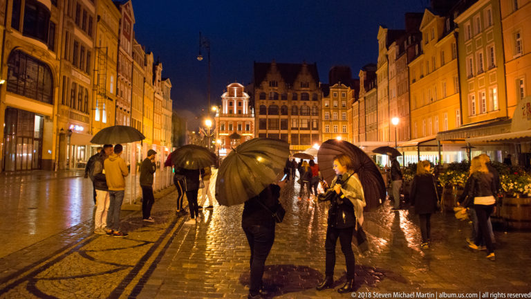 People with umbrellas stand on wet cobblestones in Wrocław Market Square beneath illuminated gabled façades.