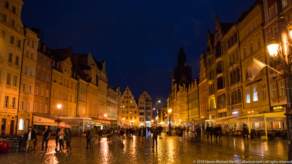 Pedestrians cross rain-wet Wrocław Market Square between illuminated historic townhouses on a blue-hour evening.
