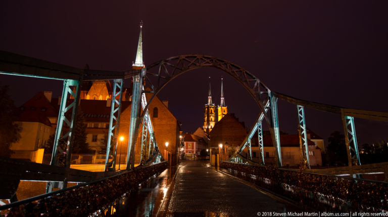 Iron Tumski Bridge at night with padlocks on the railings and church towers glowing beyond the wet deck.