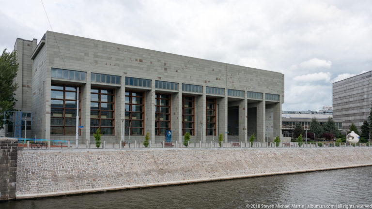 Large pale modern institutional building with tall recessed window bays facing the river embankment.