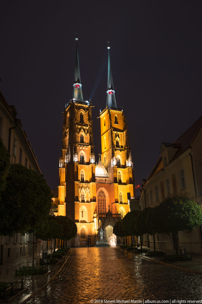 Wrocław Cathedral glowing at the end of a wet, tree-lined street on Ostrów Tumski at night.