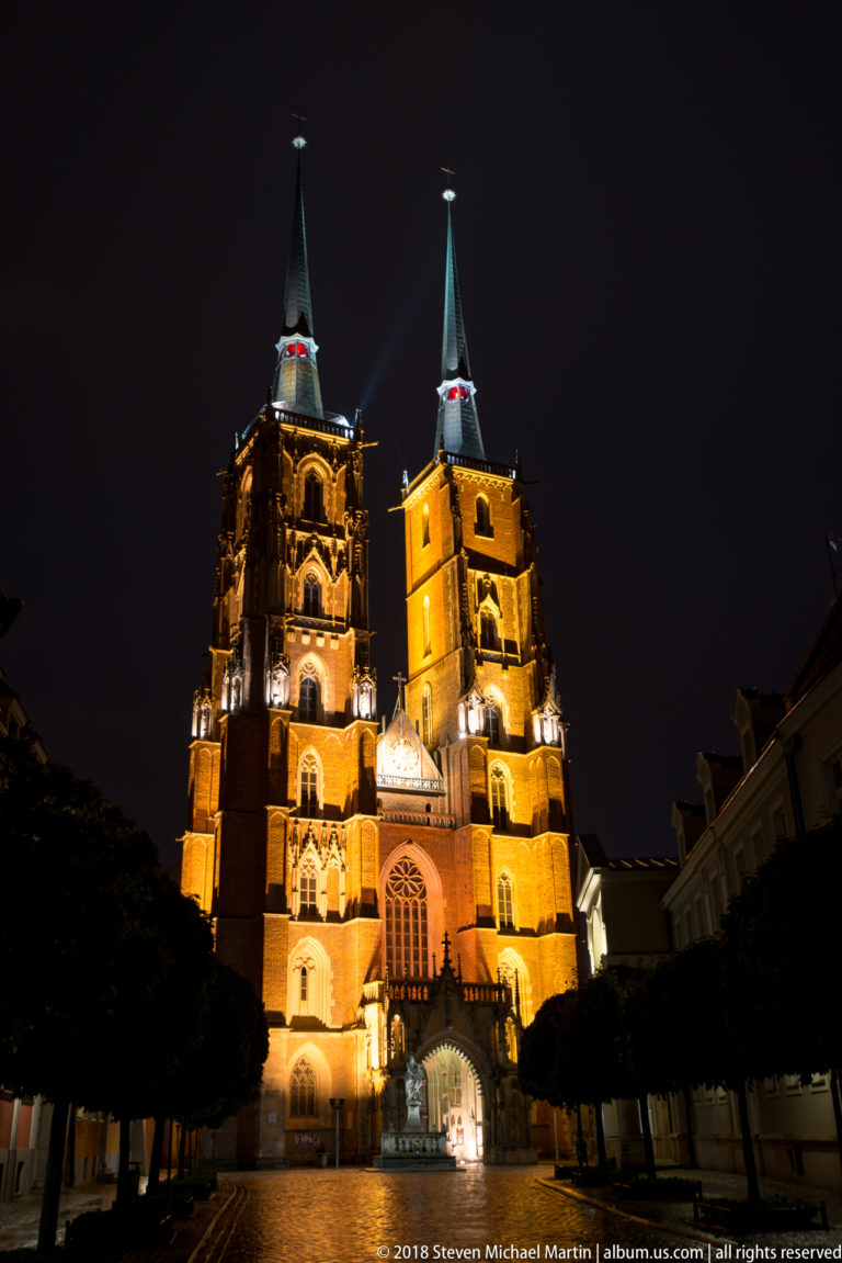 Illuminated twin-towered Gothic façade of Wrocław Cathedral at night, viewed along a tree-lined approach.