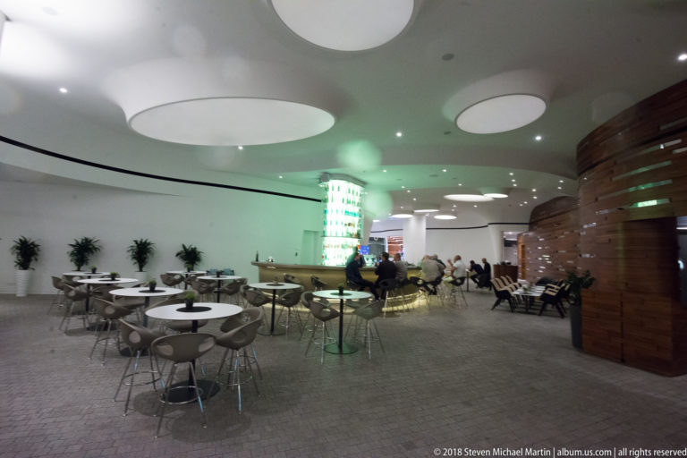Modern hotel lounge with round tables, circular ceiling panels, a curved wood wall, and guests seated in the distance.