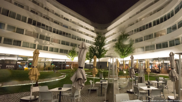 Night terrace with tables and umbrellas in the landscaped courtyard of the curved white OVO Wrocław complex.