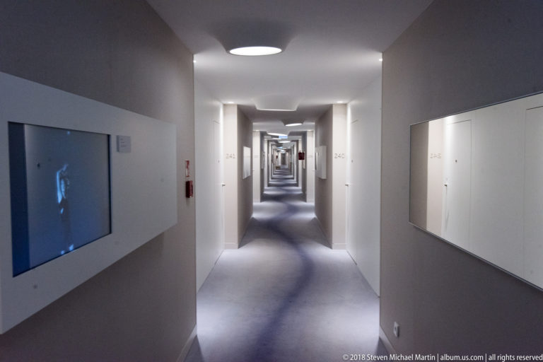 Long white hotel corridor with repeated doorways, circular ceiling lights, and a strong central vanishing point.