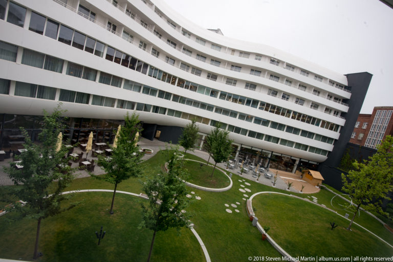 Curved white façades of OVO Wrocław surround a landscaped courtyard with grass, trees, café tables, and stepping stones.