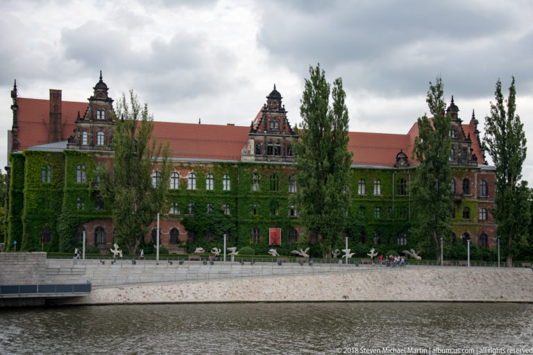 Long ivy-covered red-brick historic building with ornate gables and red roof beside the river.