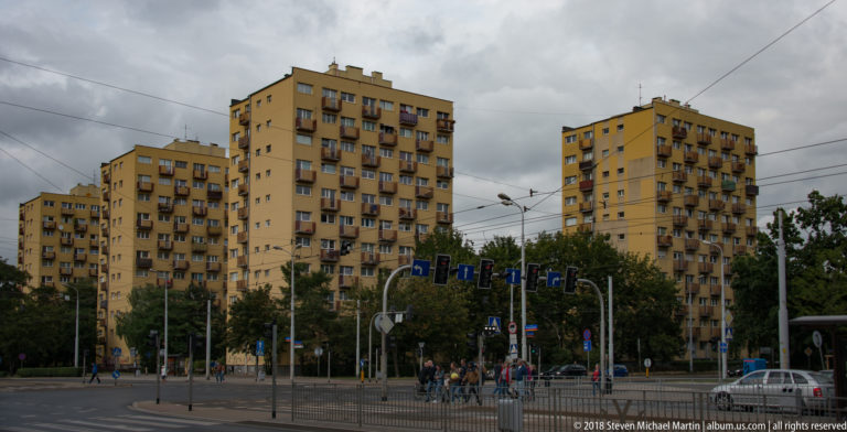 Yellow residential high-rises with balconies stand around a tram-wired city intersection under a cloudy sky.