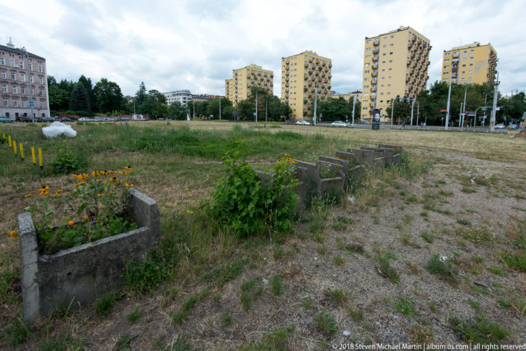 Vacant weedy lot with concrete remnants in foreground and yellow apartment towers in the background.