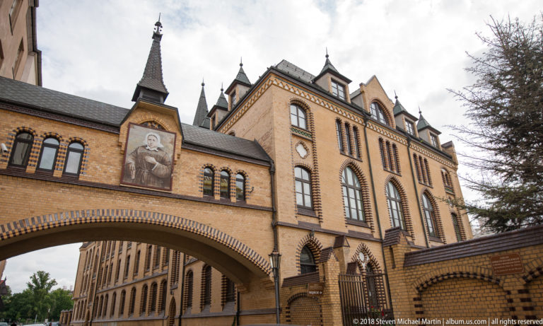 Historic brick institutional building with tall arched windows, decorative masonry, and an arched passage over the street.