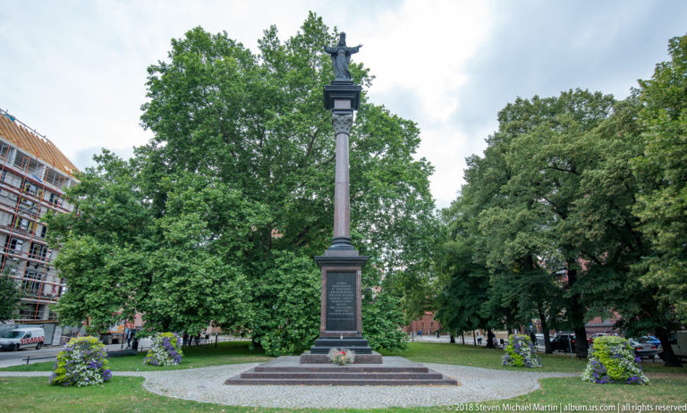 Tall stone memorial column on a stepped base in a leafy square with flowers around it.