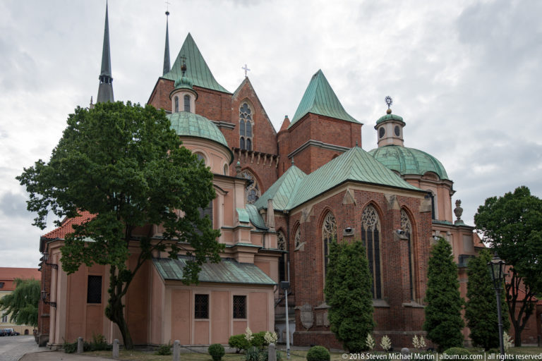 Side view of historic church with brick Gothic apses, pale stucco additions, copper roofs, and mature trees.