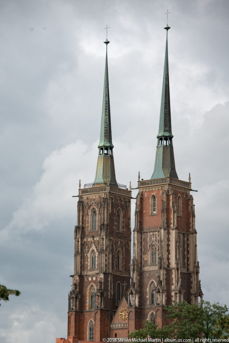 Telephoto view of Wrocław Cathedral’s twin brick towers with tall green spires against a cloudy sky.