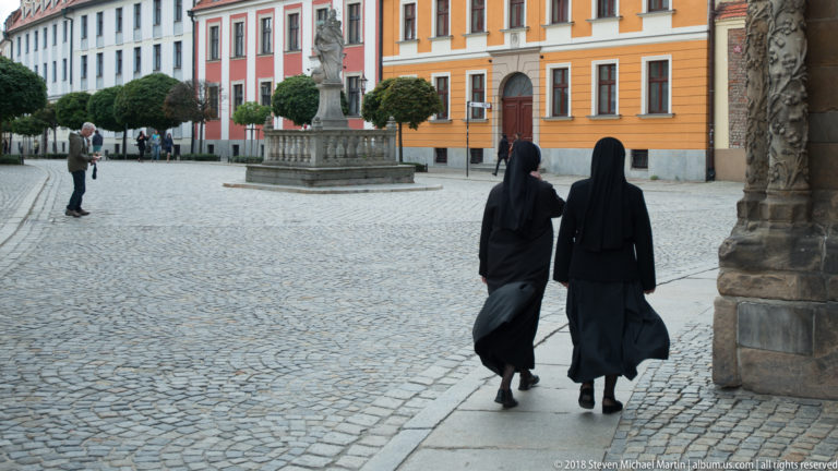 Two Catholic nuns in black habits walk across a cobbled square beside colorful historic buildings.