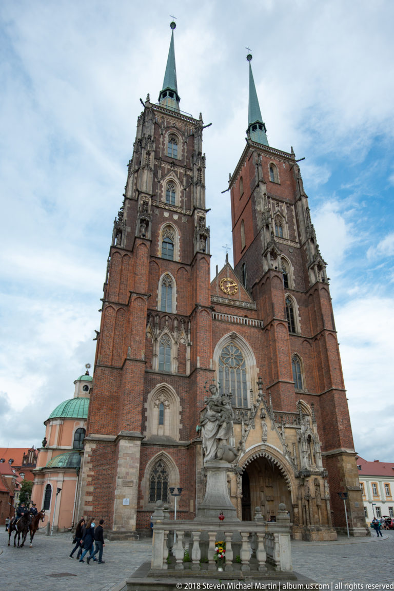 Angled view of Wrocław Cathedral with twin towers, carved entrance, and a large stone monument in the forecourt.