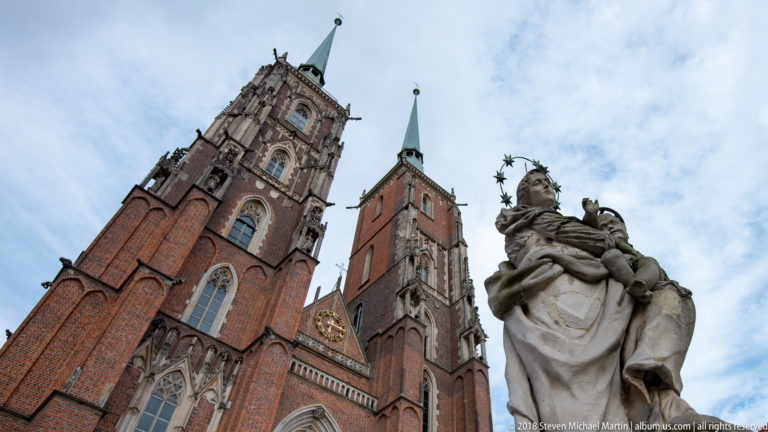 Low-angle view of Wrocław Cathedral’s twin towers behind a large stone devotional sculpture.