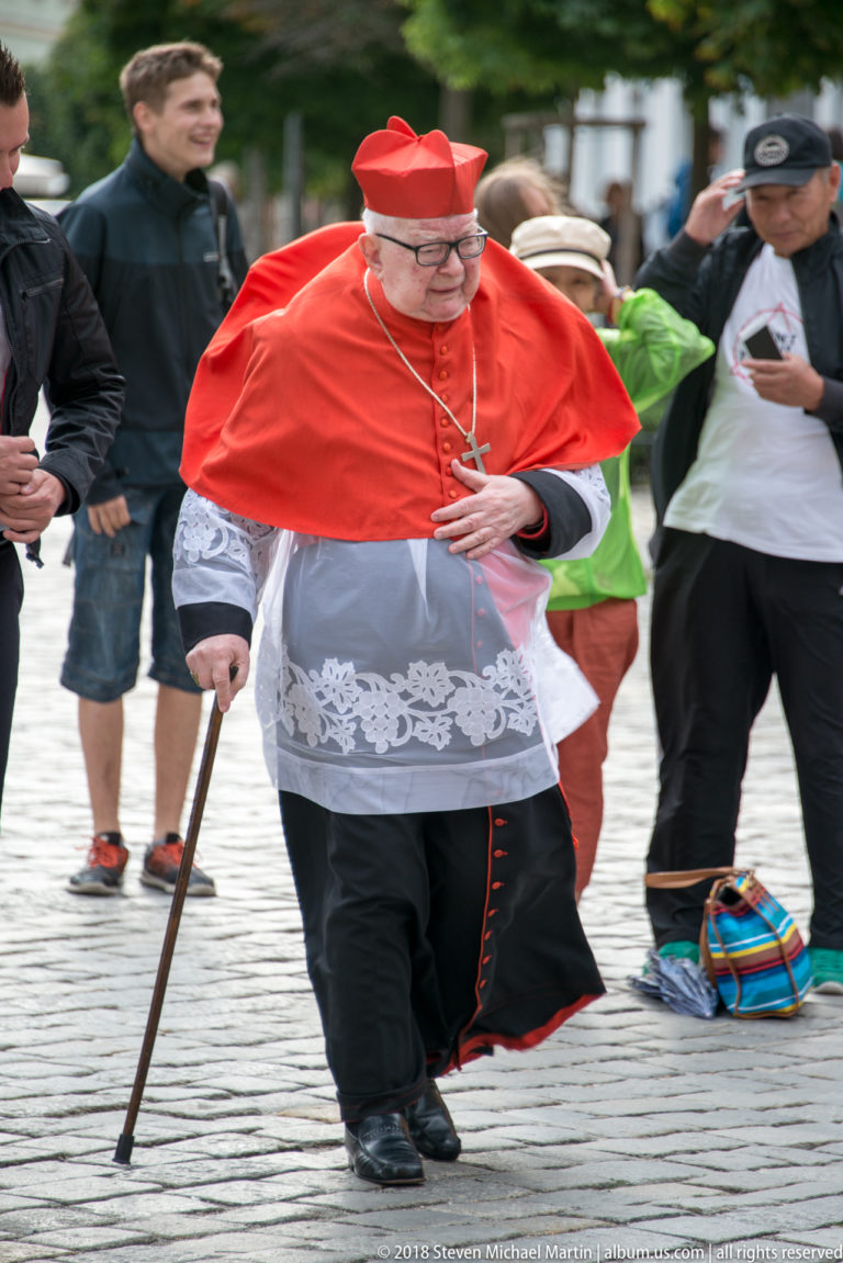 Senior Catholic cleric in red choir dress and skullcap walks with a cane on a cobbled street.